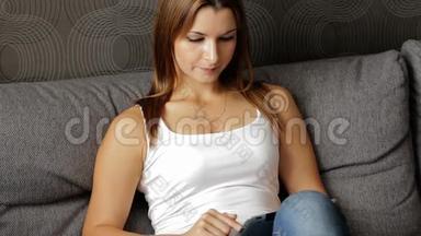 年轻微笑的女人看数字平板电脑的肖像。 有平板电脑的女人坐在沙发上。 女人用平板电脑